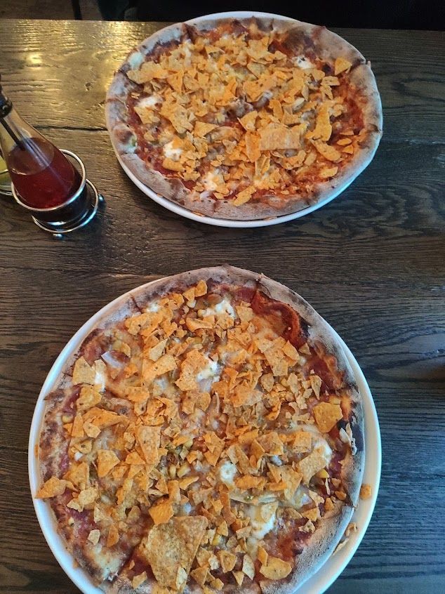 Two round pizzas topped with crushed tortilla chips, sitting on a dark wooden table next to a small bottle of hot sauce.
