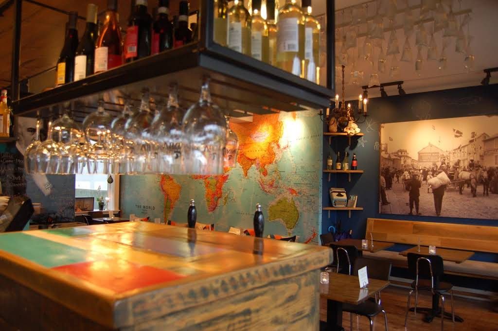 A rustic bar interior featuring a wooden counter with Italian flag colors, hanging glassware, and a world map mural.