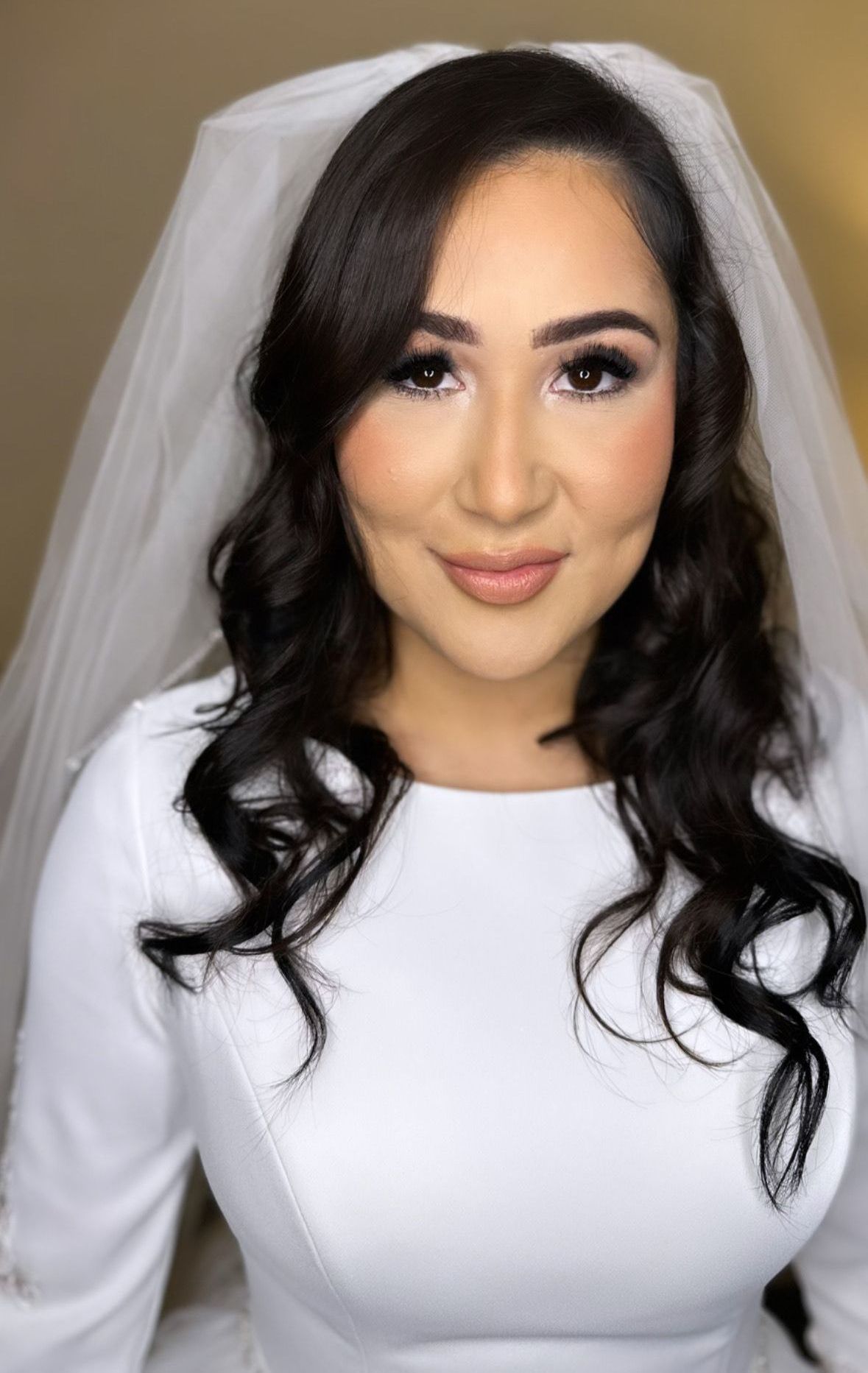 Bride in a white dress and veil, posing against a plain background with long dark curls.
