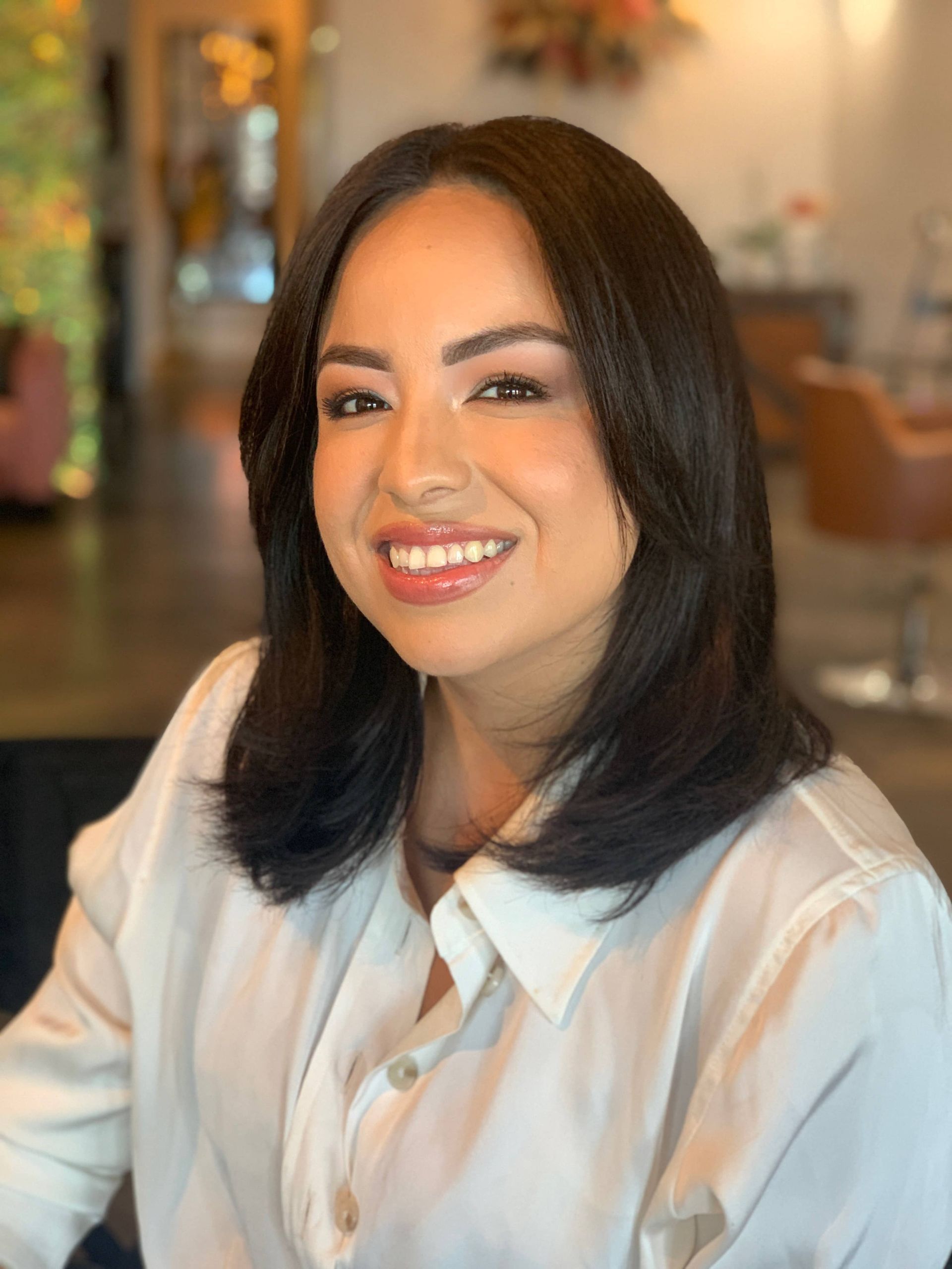 Smiling woman in a white shirt, seated indoors with warm, softly blurred background.