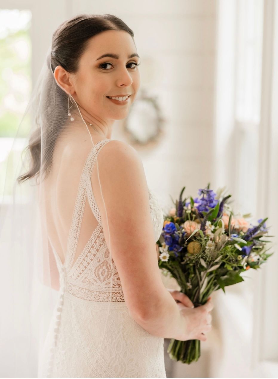 Bride in a white lace dress holding a bouquet, smiling over her shoulder near a bright window