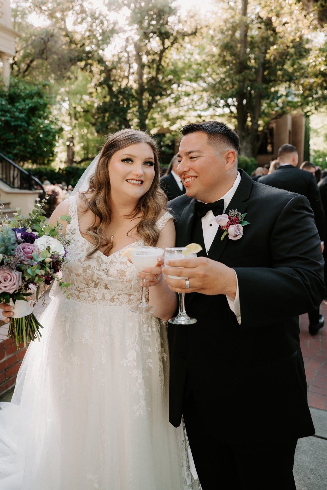 Wedding couple in formal attire toasting outdoors, smiling among flowers and greenery.