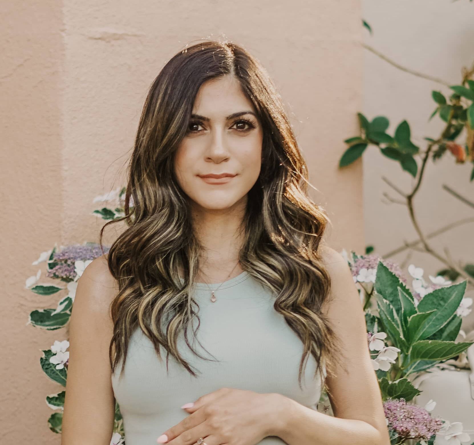 Woman with wavy hair in a light dress, standing against a pastel wall with flowers and greenery.