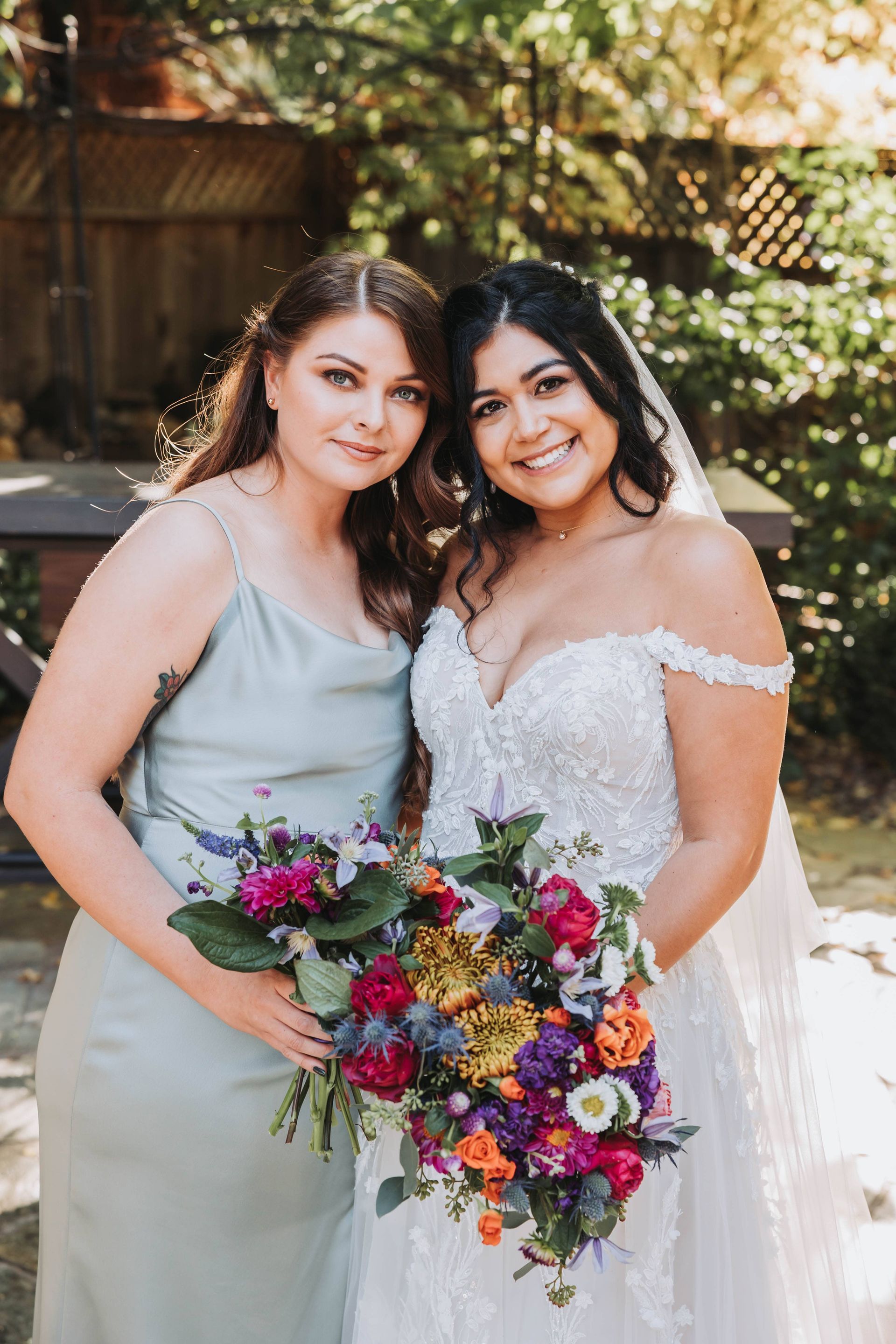Two brides in white dresses smiling and holding a colorful bouquet outdoors