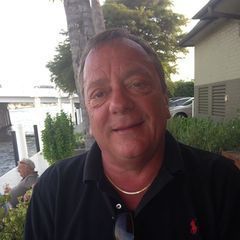 Man with short brown hair wearing a black polo shirt, gold chain, smiling outside near water.