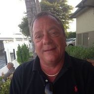 Man with short brown hair wearing a black polo shirt, gold chain, smiling outside near water.
