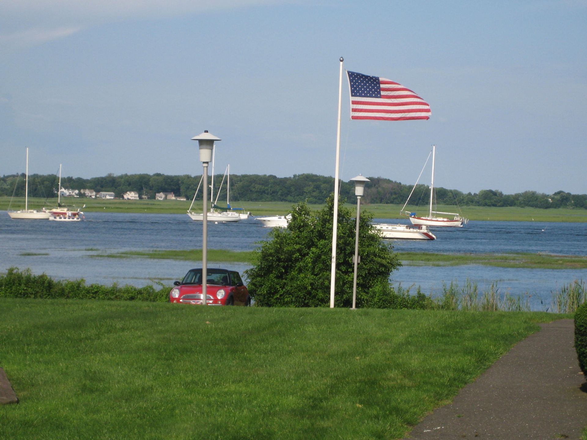 Red car near a grassy area with water and sailboats, American flag waving in the wind.