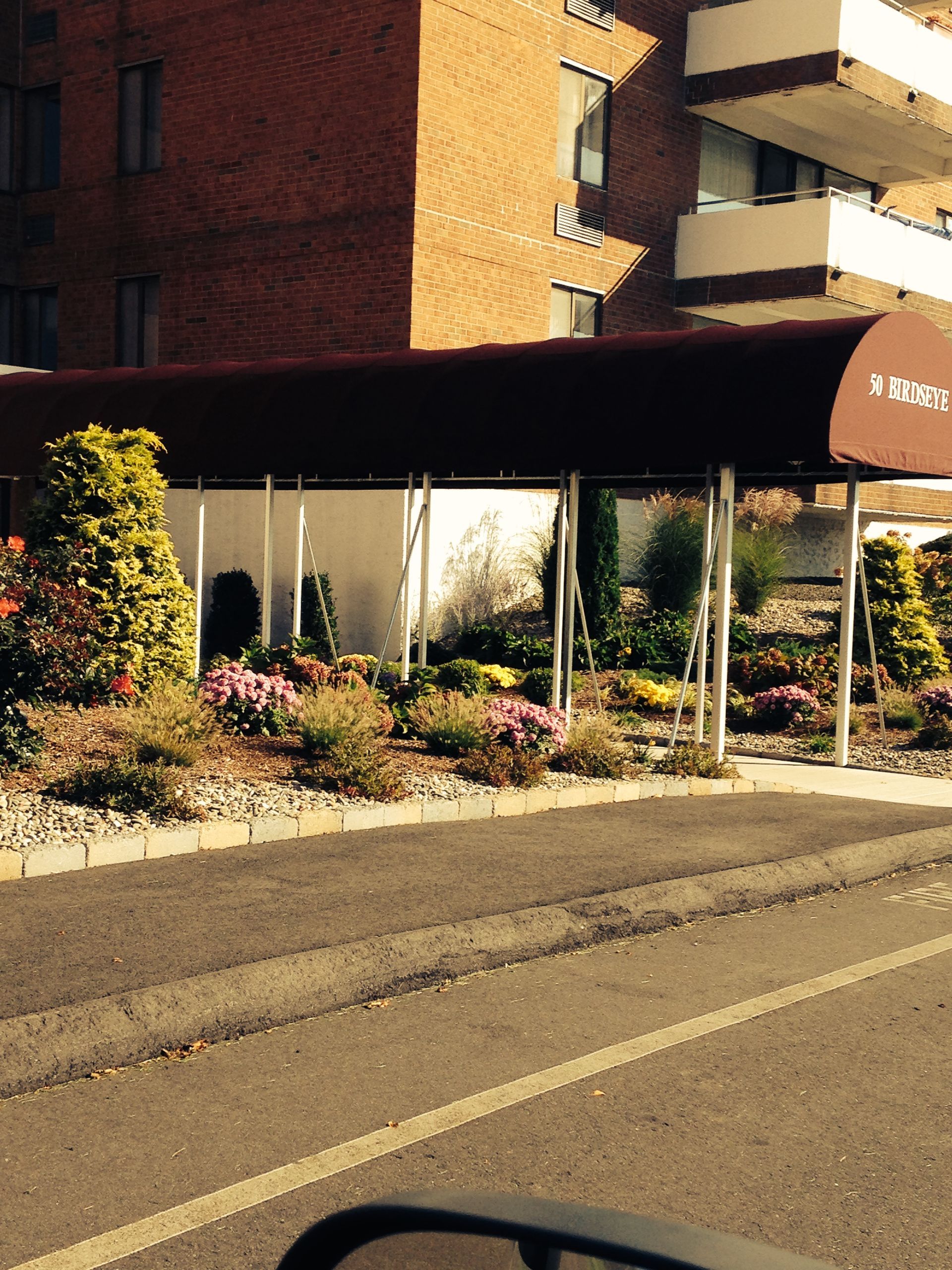 Apartment building exterior with a covered entrance, flowerbeds, and a street.