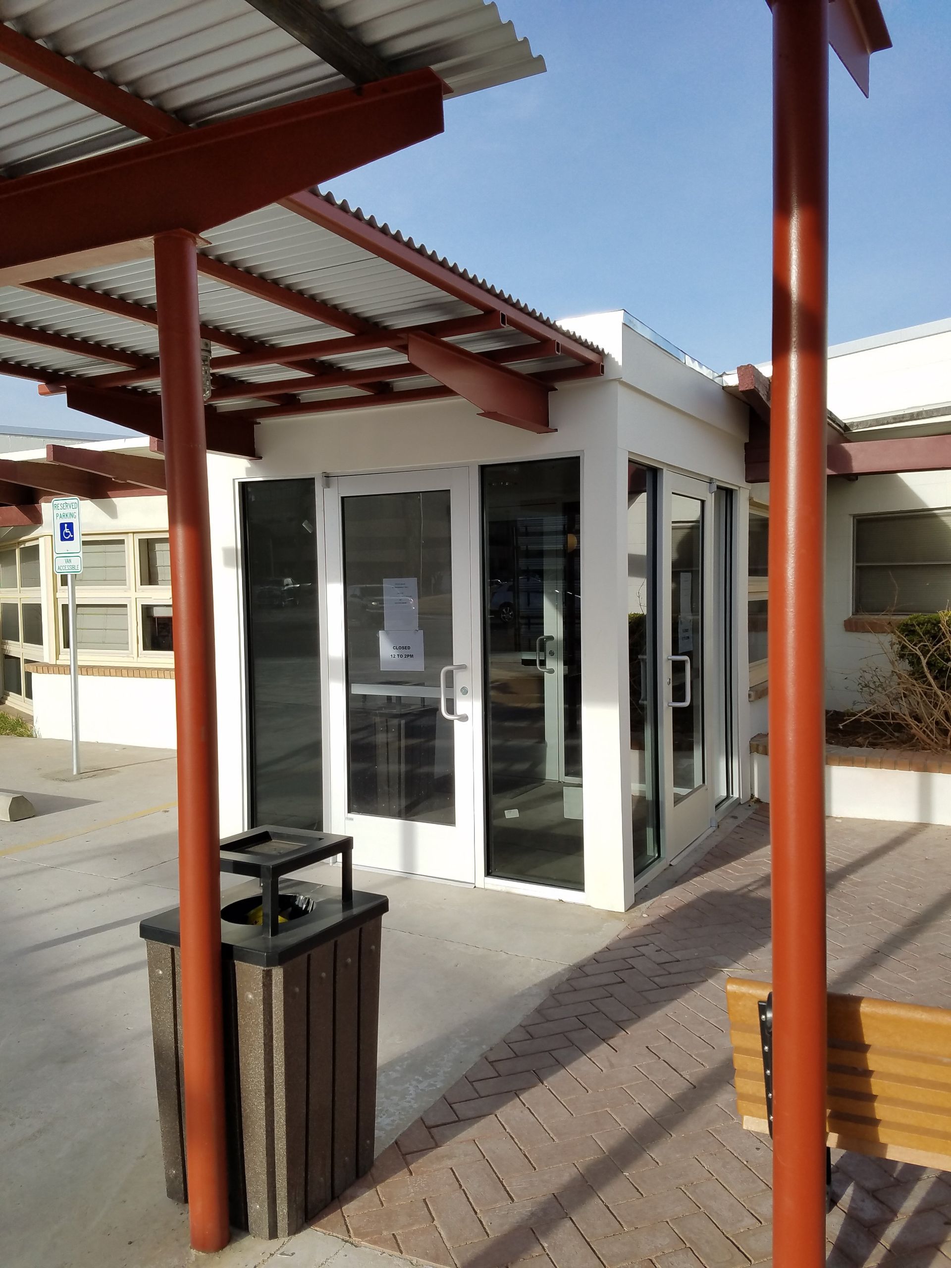 Small white kiosk with glass doors under a red-roofed shelter beside a walkway and bench