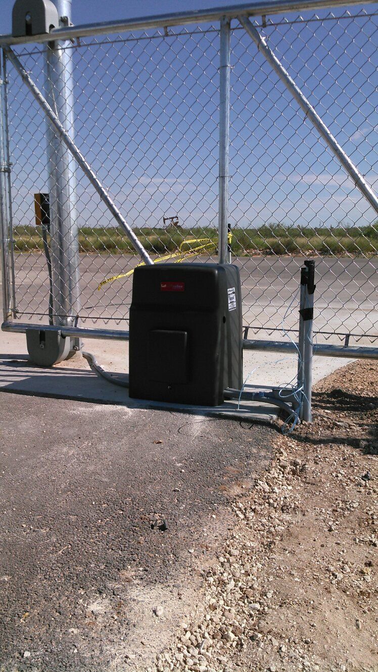 Black gate motor at the base of a chain-link sliding gate on a gravel driveway under a clear sky