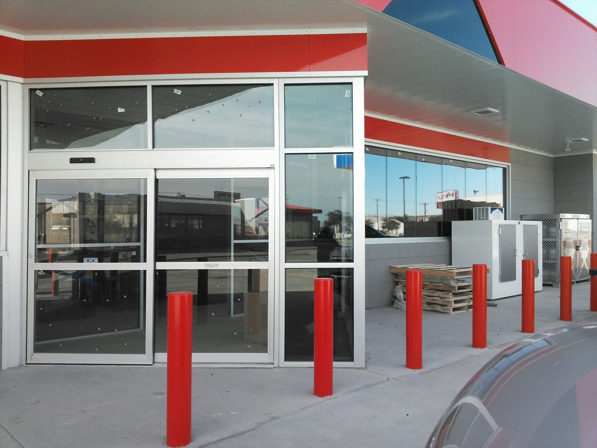Storefront entrance with glass doors, red bollards, and a covered curbside pickup area