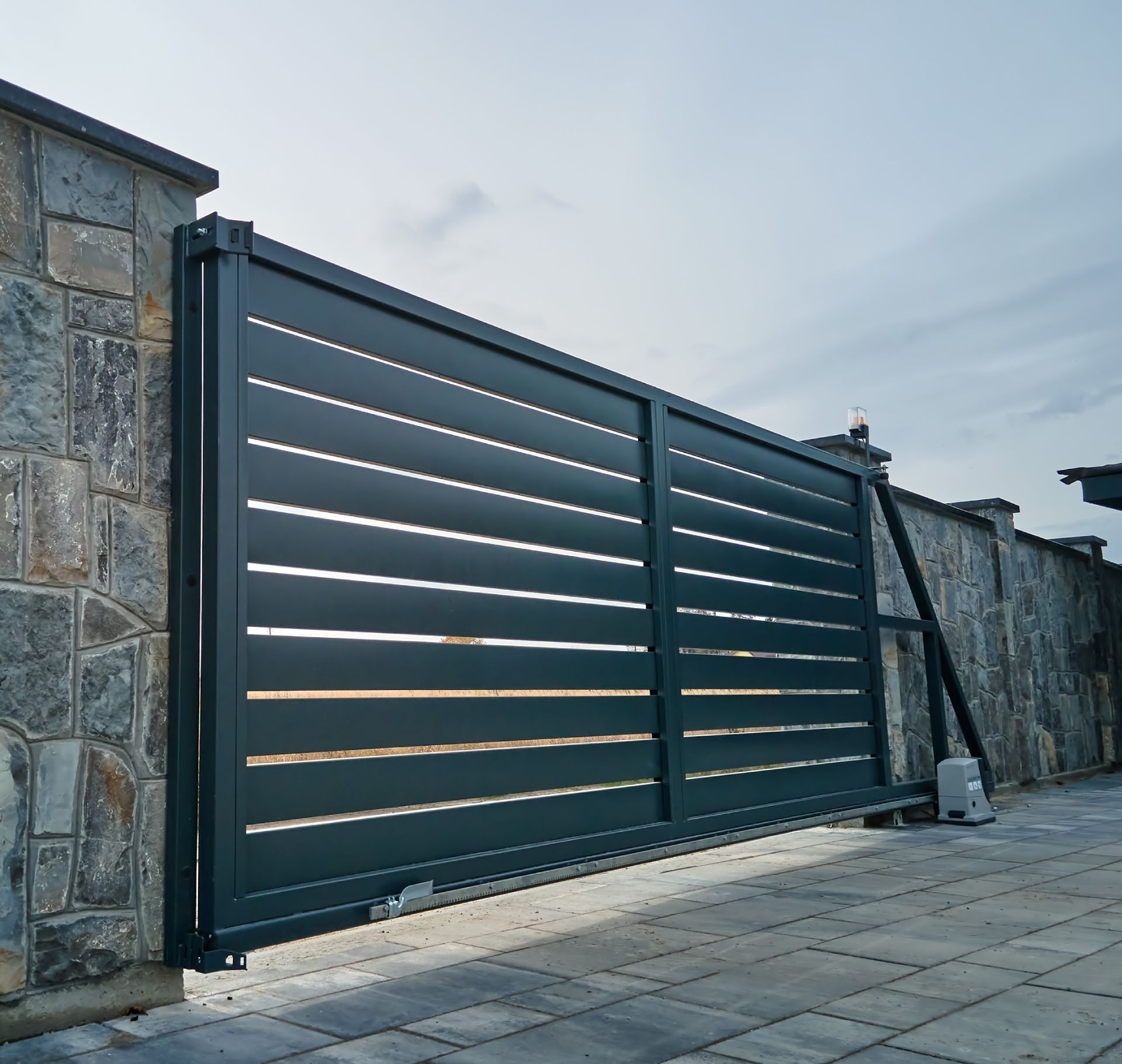Dark gray slatted metal driveway gate beside a stone wall on a paved walkway