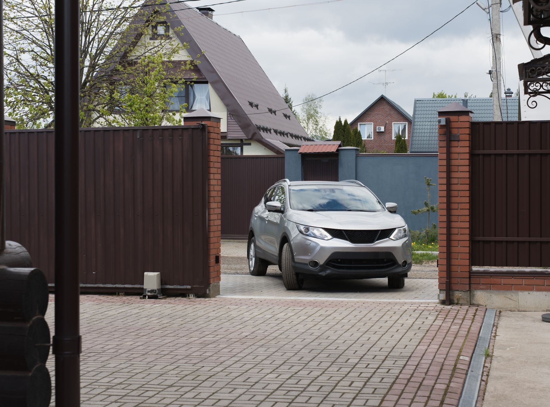 Silver SUV parked in a driveway between brown gates, with houses and trees in the background.