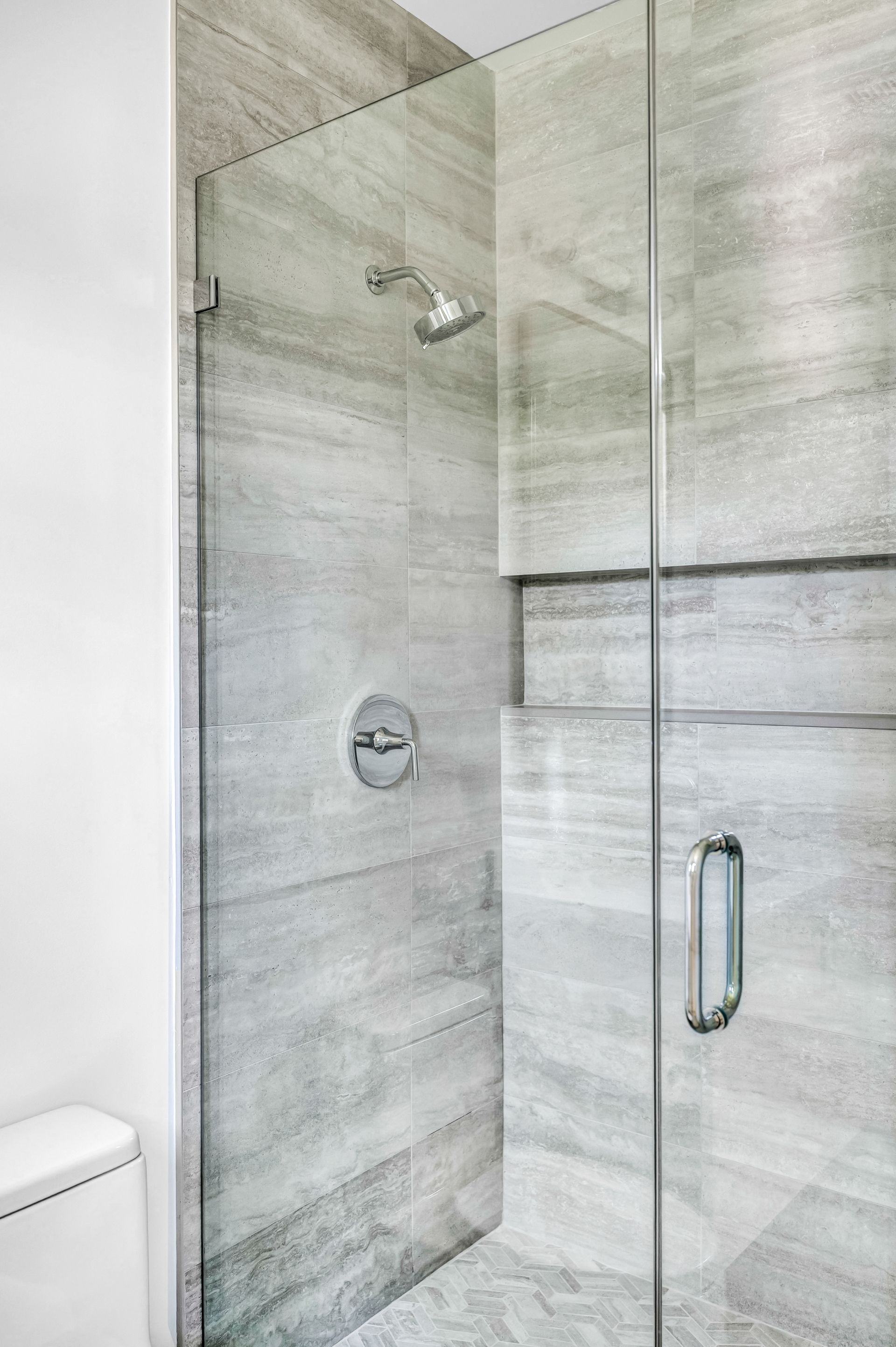 Glass shower enclosure in a light gray tiled bathroom, with built-in shelves and a chrome handle.
