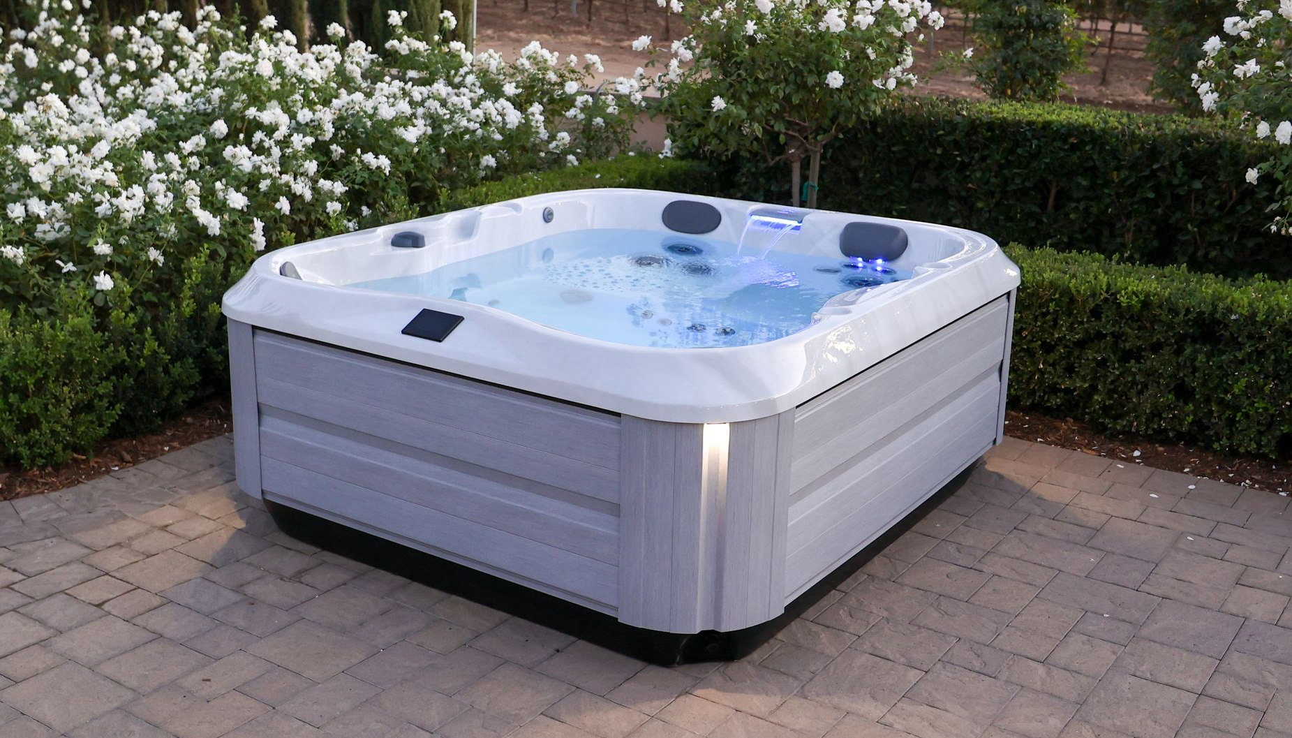 White outdoor hot tub with blue water on a stone patio, surrounded by hedges and flowers