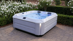 White outdoor hot tub with blue water on a stone patio, surrounded by hedges and flowers