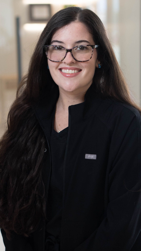 Zenia wearing glasses and a brown jacket is smiling for the camera.