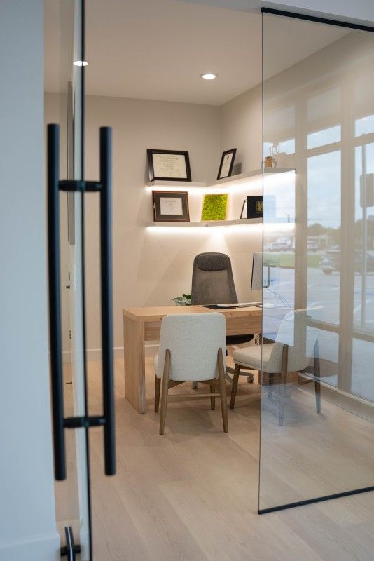 Small modern office with a wooden desk, white chair, shelves, and glass walls viewed through an open doorway - Transcend Dental Office Photo