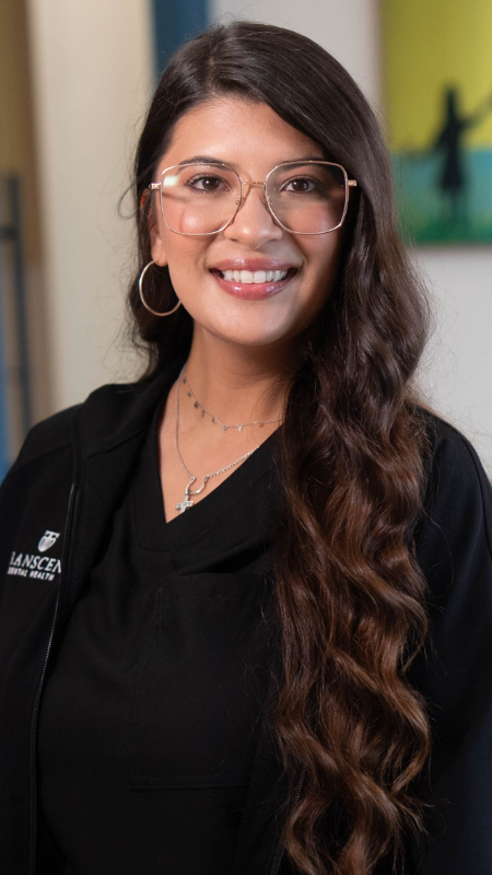Zenia wearing glasses and a brown jacket is smiling for the camera.