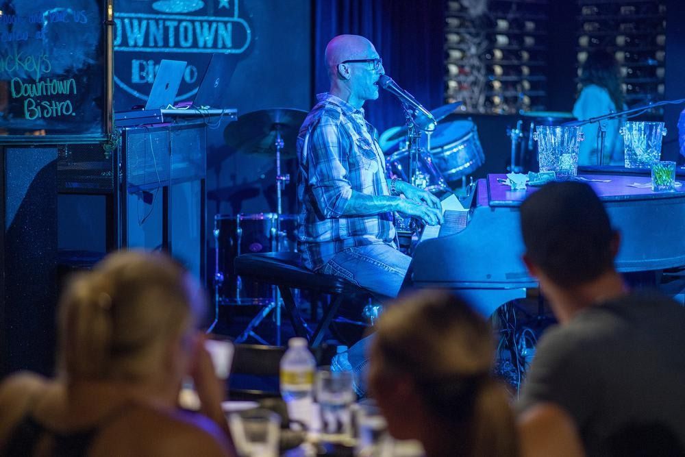 Blue-lit musician playing drums on stage in a bar, with blurred audience in the foreground