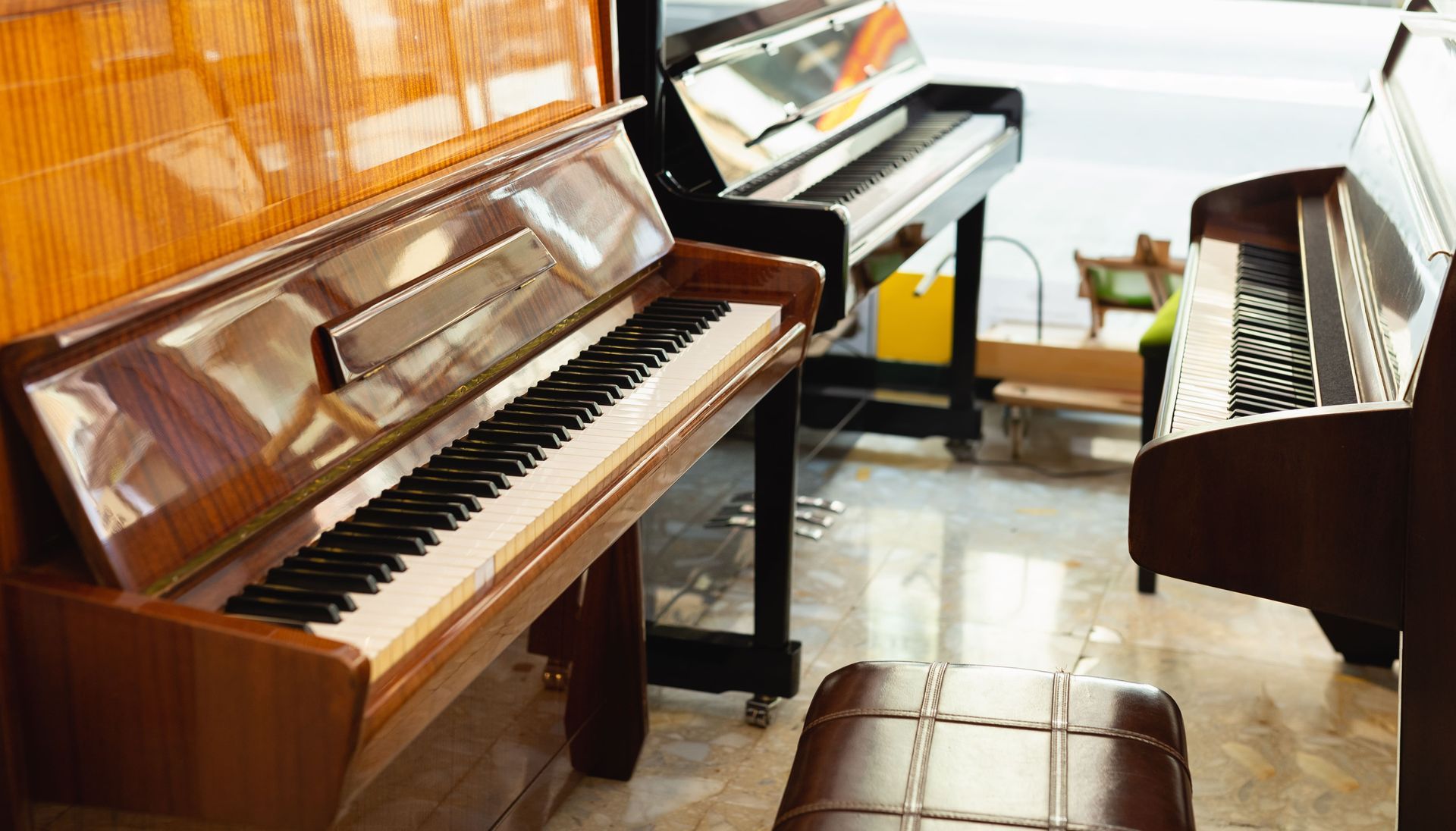 Several upright pianos in a bright room with a window and bench