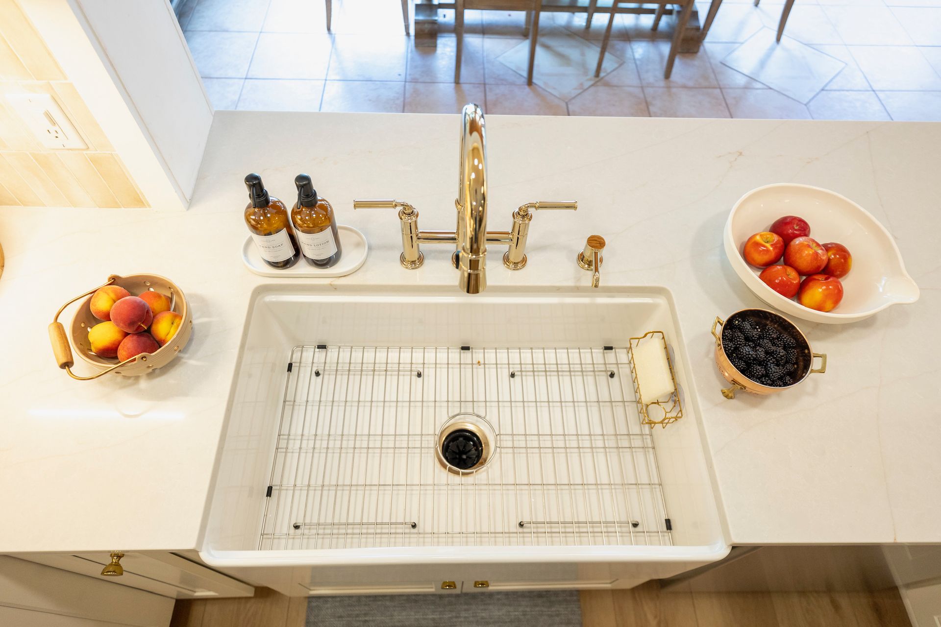 Top-down view of a bright kitchen sink with gold faucet, fruit bowls, soap bottles, and a wire drying rack. - BittnerSweet Co.