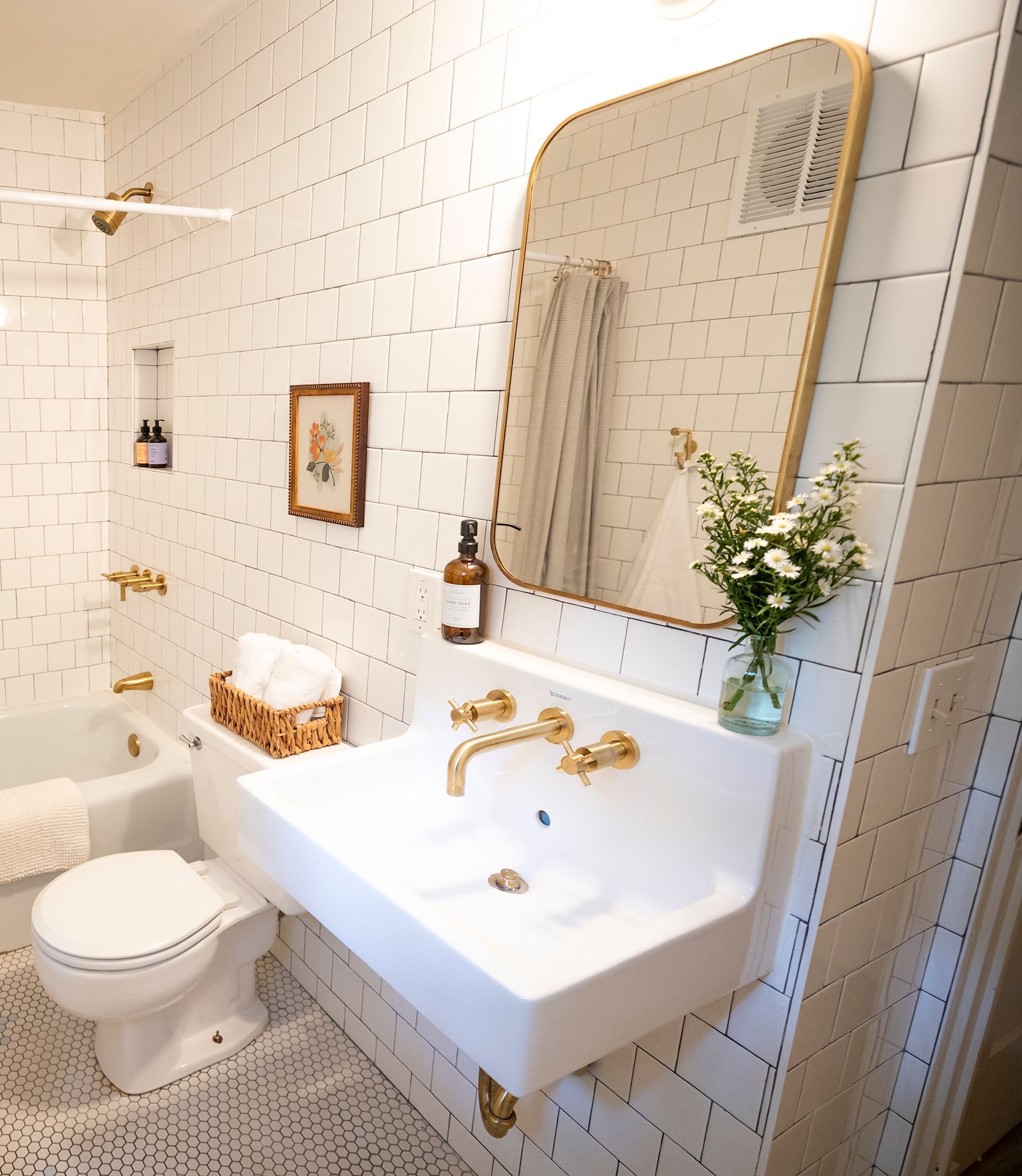 Bright white tiled bathroom with a wall-mounted sink, toilet, gold fixtures, mirror, and flowers - BittnerSweet Co.