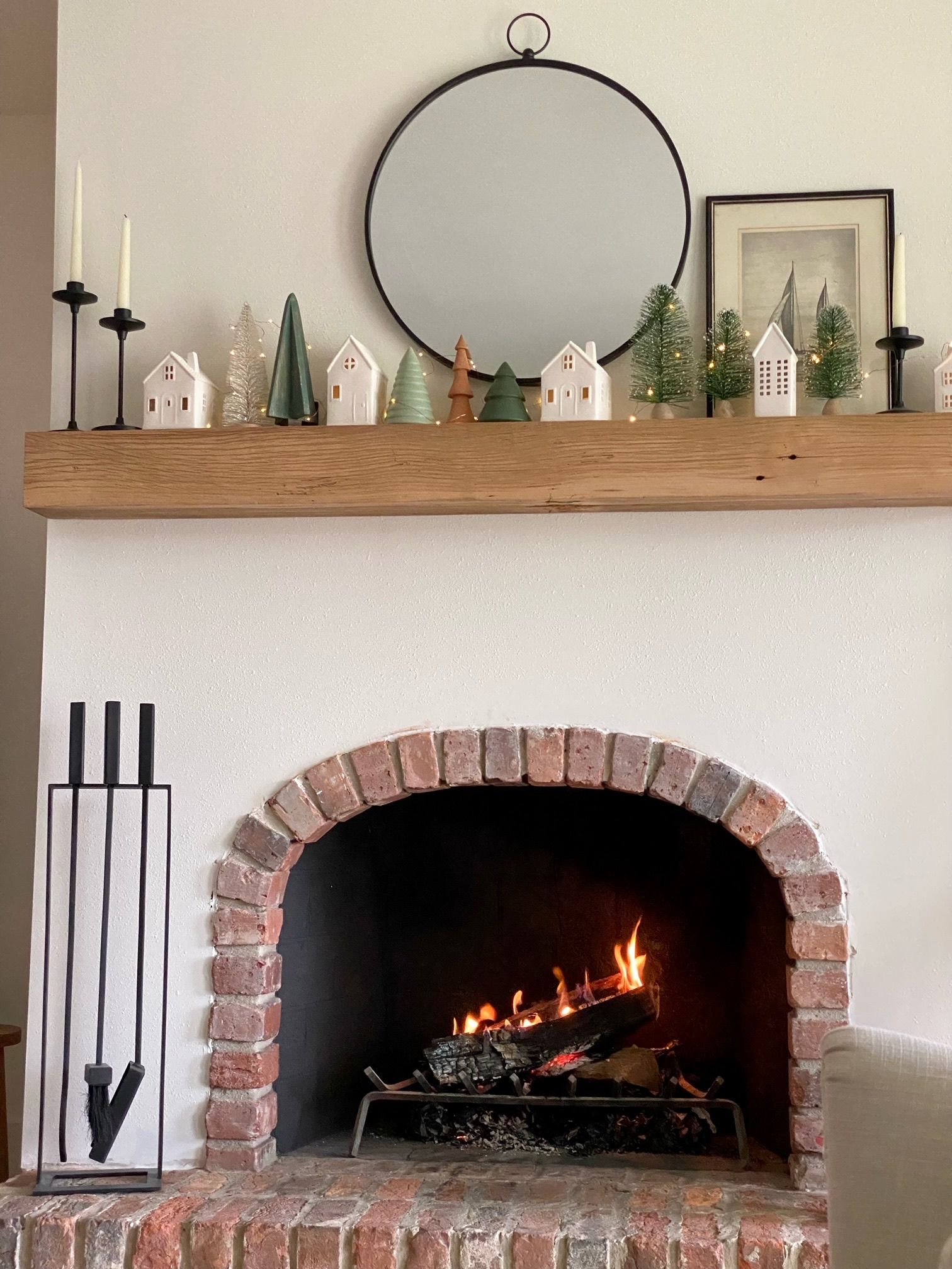 Cozy fireplace with a fire burning, brick arch, and decorated mantel with mirror and small trees - BittnerSweet Co