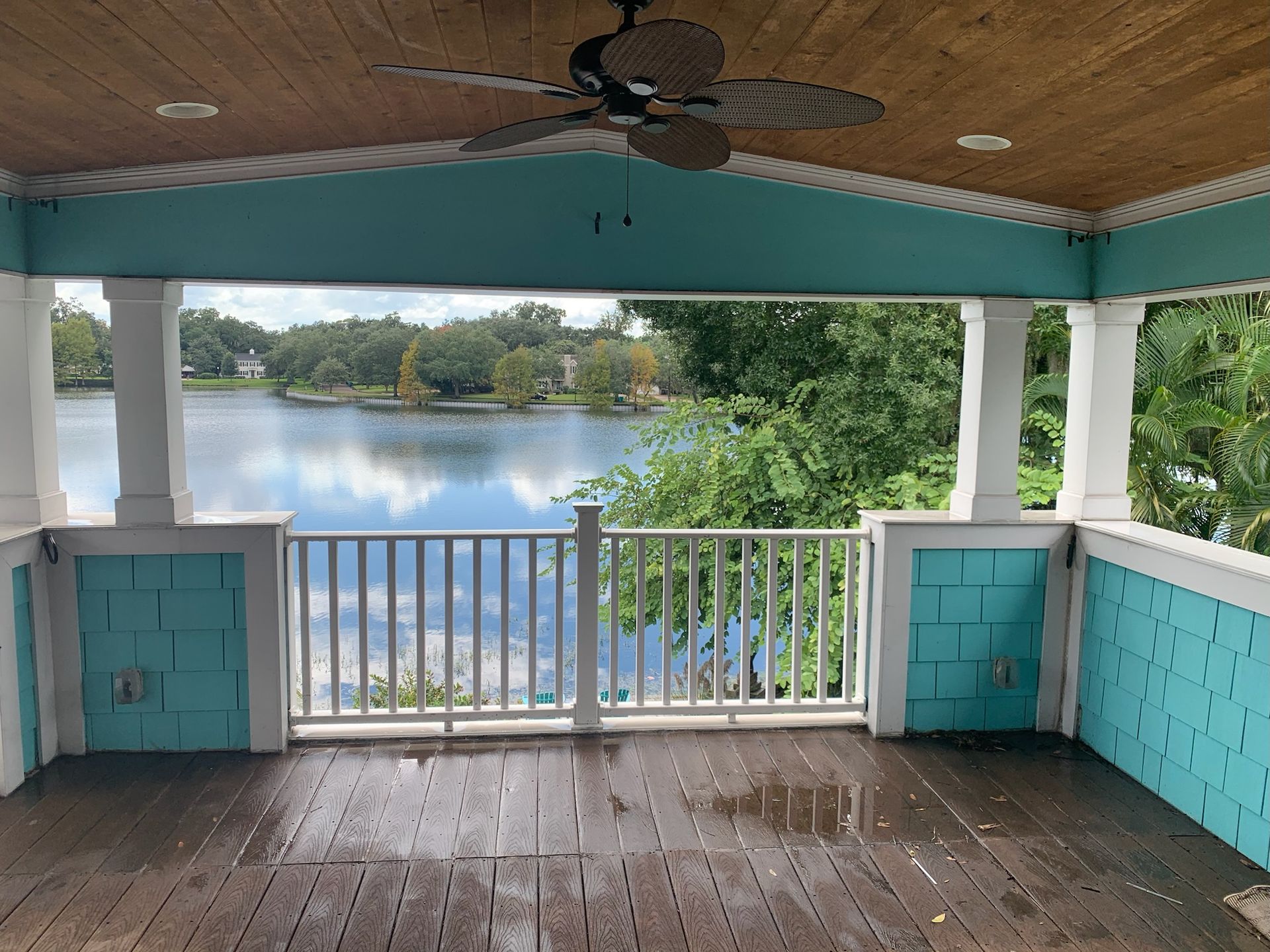 Covered porch with turquoise railings overlooking a calm lake and trees, with a ceiling fan overhead - BittnerSweet Co.