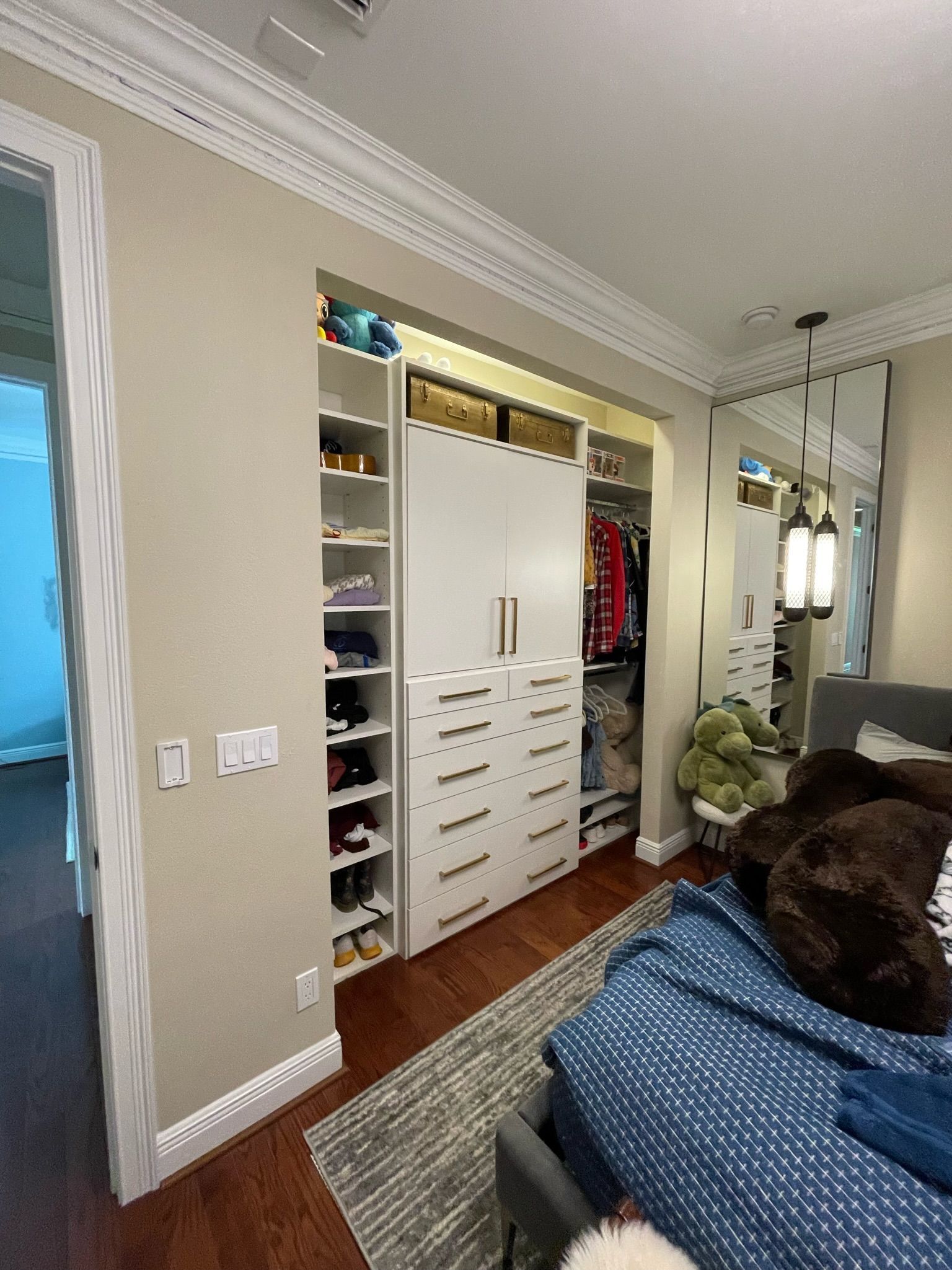 Bedroom with open white closet, hanging clothes, and bed with blue comforter beside a window - BIttnerSweet Co.