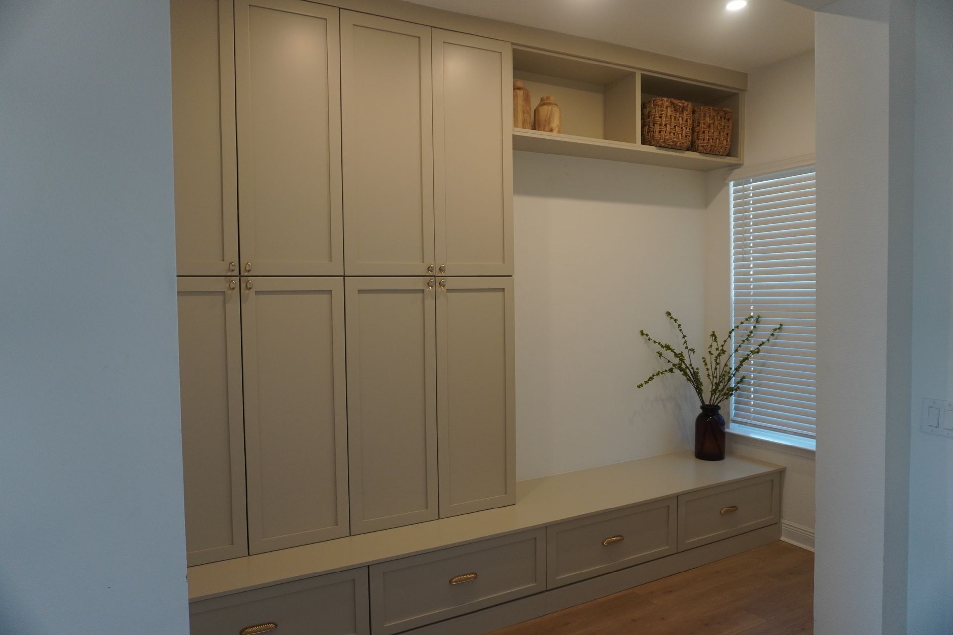 Built-in beige cabinets and drawers in a bright room, with a window, shelves, and a vase of flowers. - BittnerScweet Co