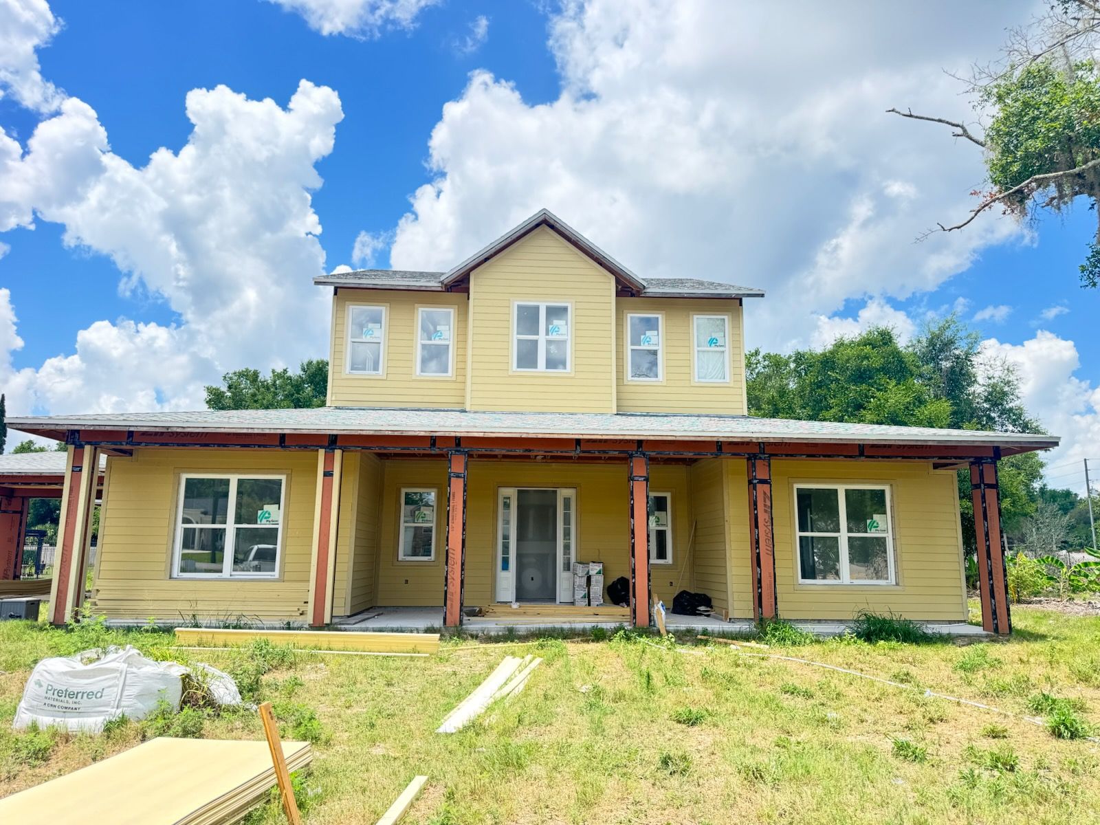 Yellow two-story house with a wraparound porch, white trim, and a grassy front yard under a blue sky.