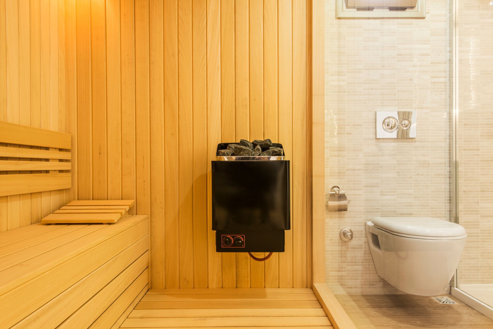 BittnerSweet's wood-paneled sauna with benches, heater, and a toilet beside a glass shower enclosure.