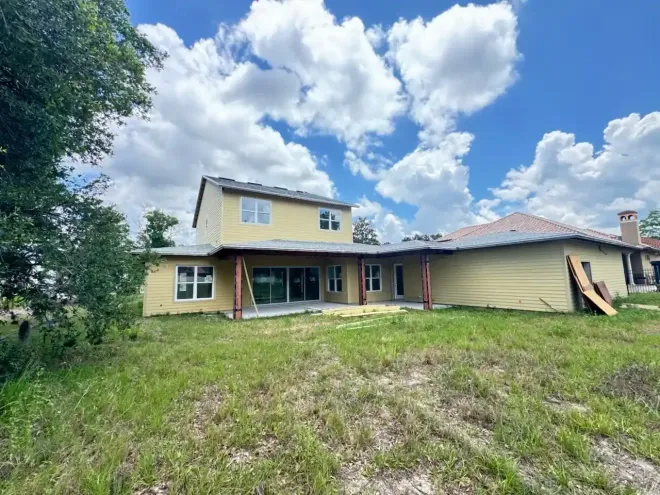 Overgrown backyard with a yellow two-story house and covered porch under a cloudy sky
