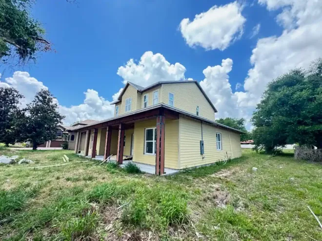 Yellow two-story house with a porch, red trim, and a grassy yard under a blue sky with clouds.