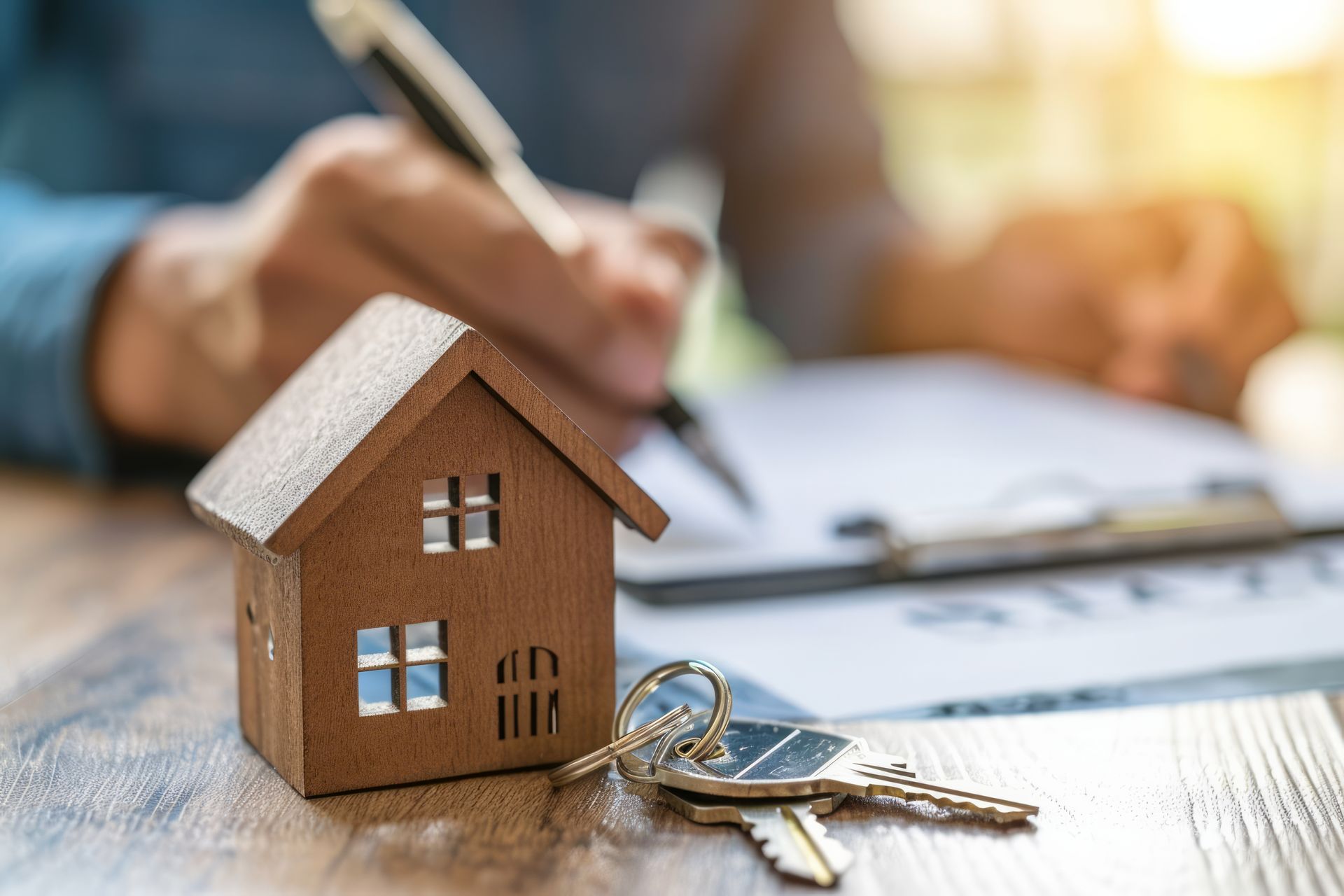 A wooden house model and a set of keys sit on a desk in front of a person signing a document.