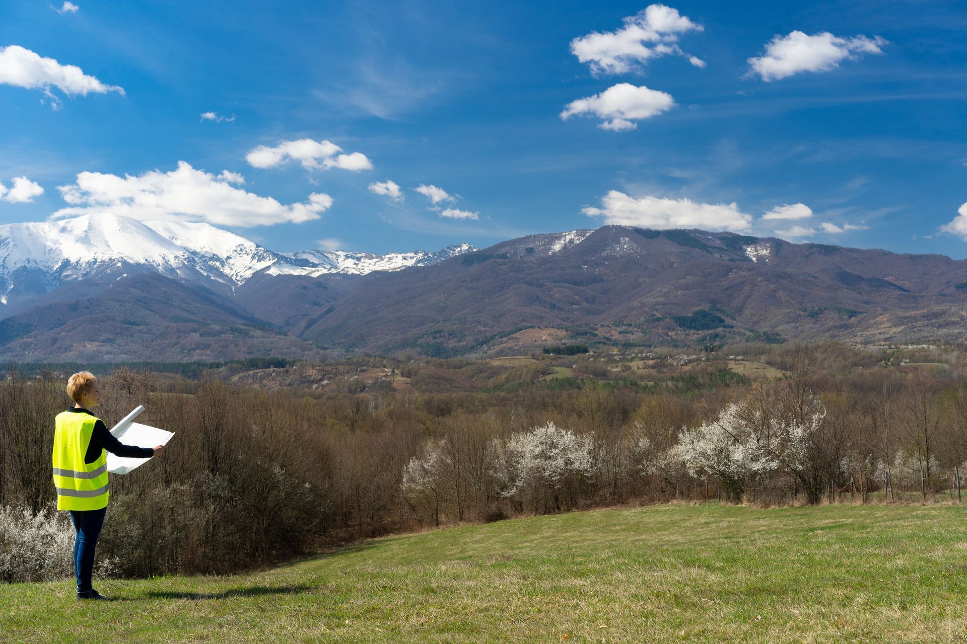 A person in a high-visibility vest stands in a grassy field holding plans, looking toward snow-capped mountains.