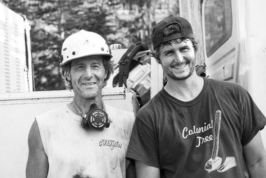 A black-and-white portrait of two individuals standing side by side, one wearing a hard hat and respirator, the other a cap.