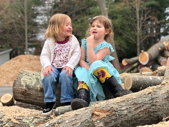 Two children sit on a pile of logs in a woodsy setting, smiling and chatting.