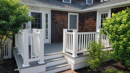 A backyard deck with white railings and stairs, connected to a house with brown shingle siding and green bushes.