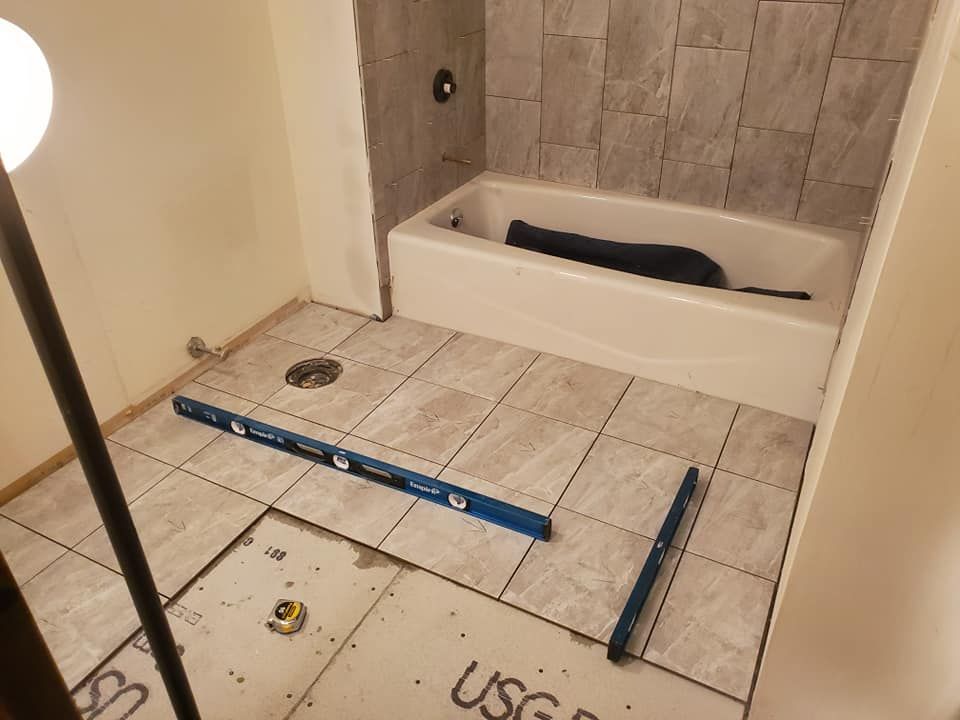 A bathroom under construction showing grey tile flooring, a bathtub, and two levels placed on the floor for installation.
