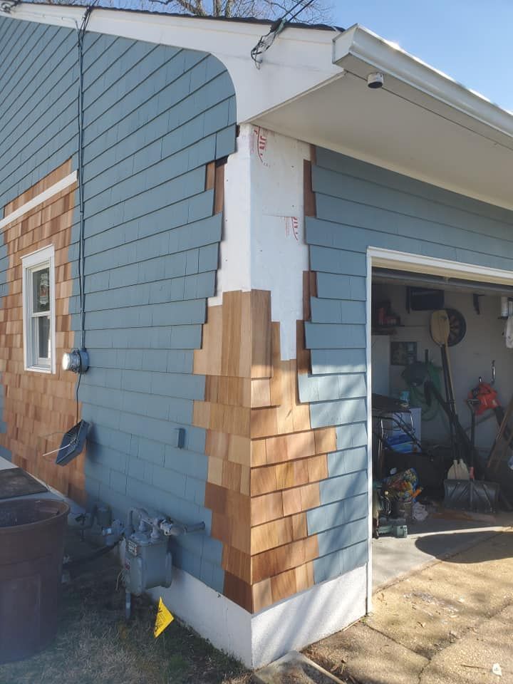 A house exterior corner showing unfinished cedar shingles against partially painted blue siding on a sunny day.