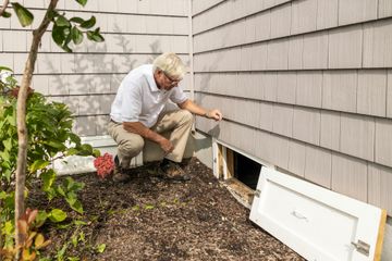 A person kneels in mulch, inspecting a crawl space opening in the side of a house with grey wood-shingle siding.