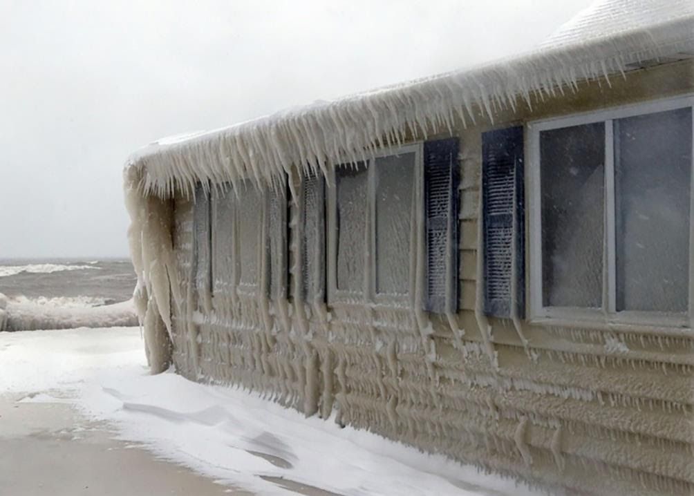 A seaside house completely encased in thick, freezing ice, with icicles hanging from the roof and windows.