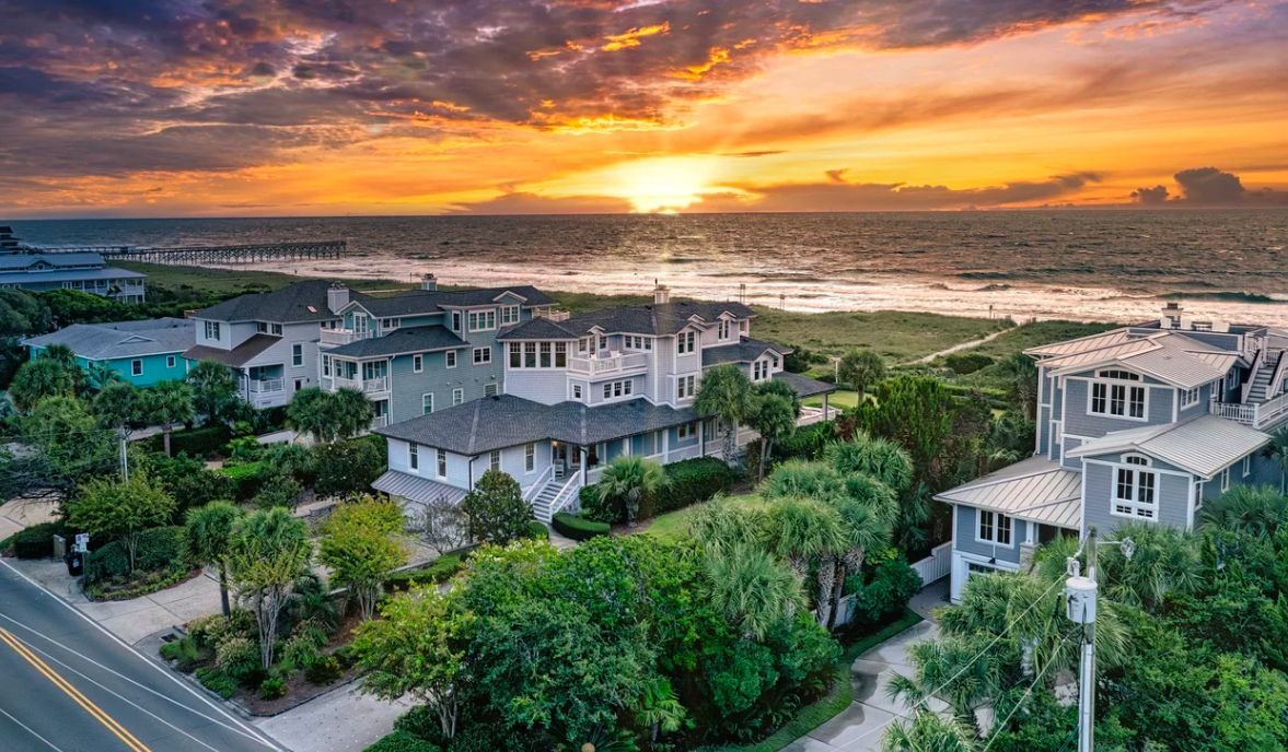 An aerial view of coastal homes nestled among green trees facing the ocean at sunset with a golden, cloudy sky.