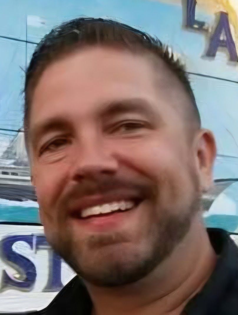 A smiling person with a groomed beard and short dark hair, shown in a close-up against an outdoor background.