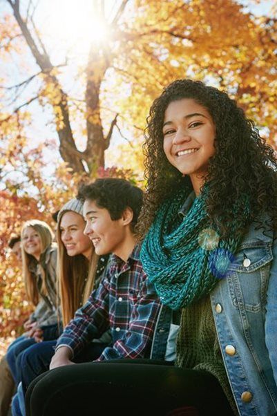 Four people smiling while sitting together outdoors in an autumn forest with bright, sunlit yellow leaves.