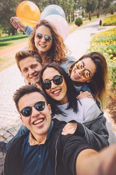 A group of five friends wearing sunglasses smiles while taking a group selfie outdoors with balloons in the background.