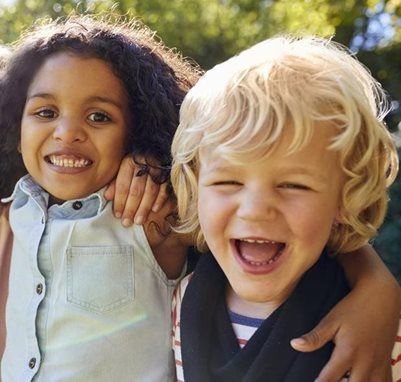 Two children smiling, with one arm resting around the other's shoulder, outdoors in soft, natural light.