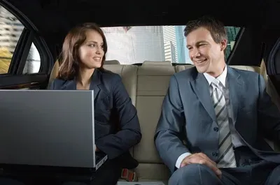 Woman and man in suits smile at each other in the back of a car; she uses a laptop.